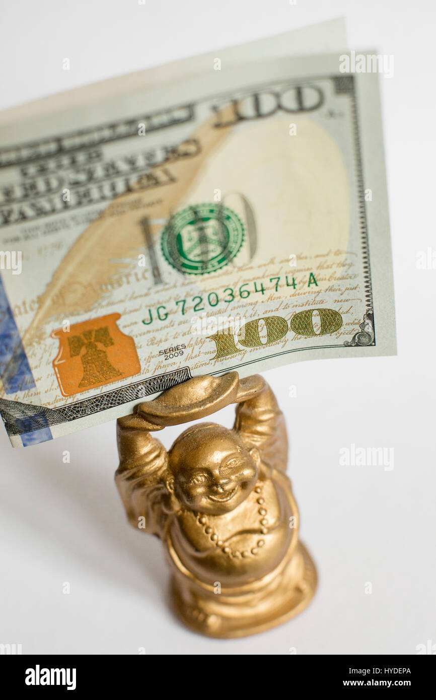 a small golden buddha figurine standing on a white back ground holds a one hundred dollar bill in United States currency above his head Stock Photo