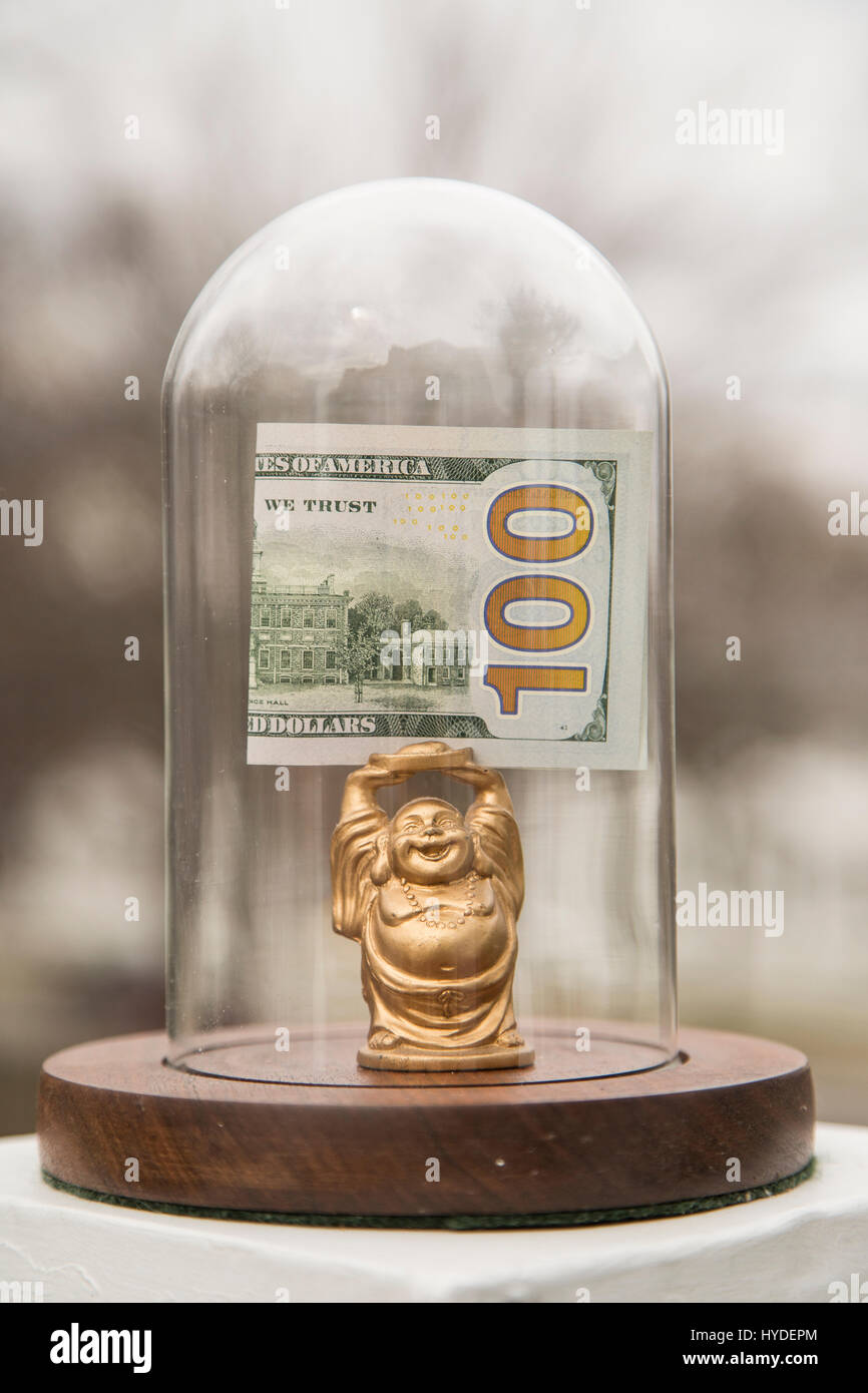 a small golden buddha figurine standing in a glass display dome holds a folded one hundred dollar bill above his head Stock Photo