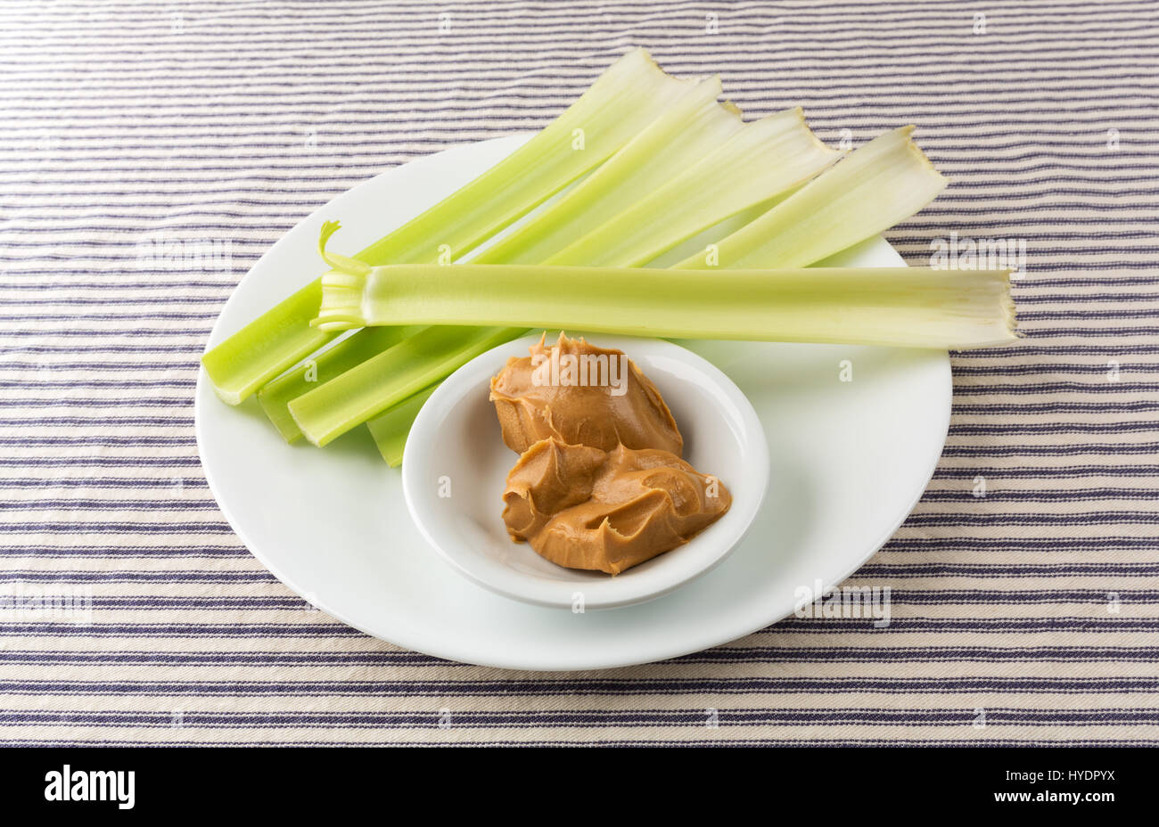 Several celery stalks with a bowl of peanut butter on a white plate atop a blue striped tablecloth. Stock Photo