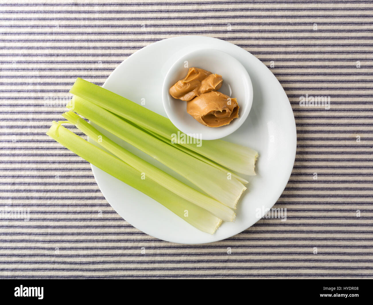 Top view of several celery stalks with a bowl of peanut butter on a white plate atop a blue striped tablecloth. Stock Photo