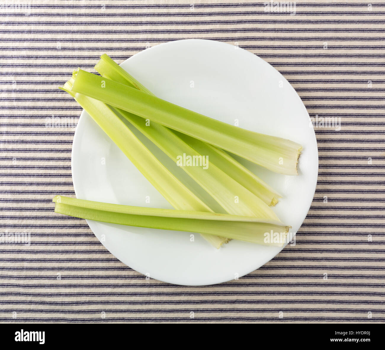 Top view of several celery stalks on a white plate atop a blue striped tablecloth. Stock Photo