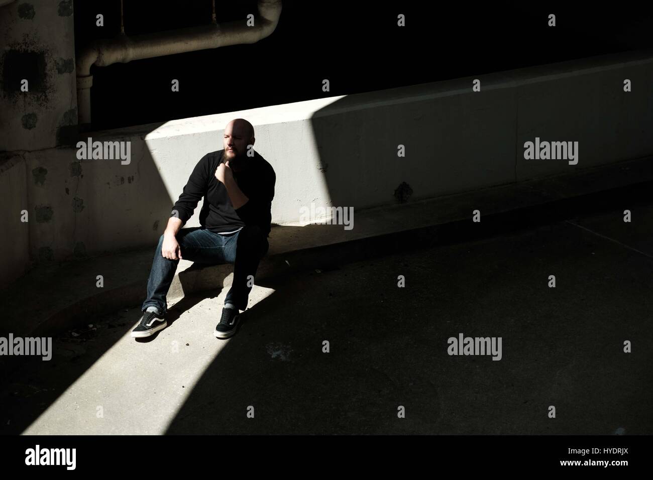 Portrait of a man with bald head and long beard sitting in a sliver of light in a parking garage Stock Photo