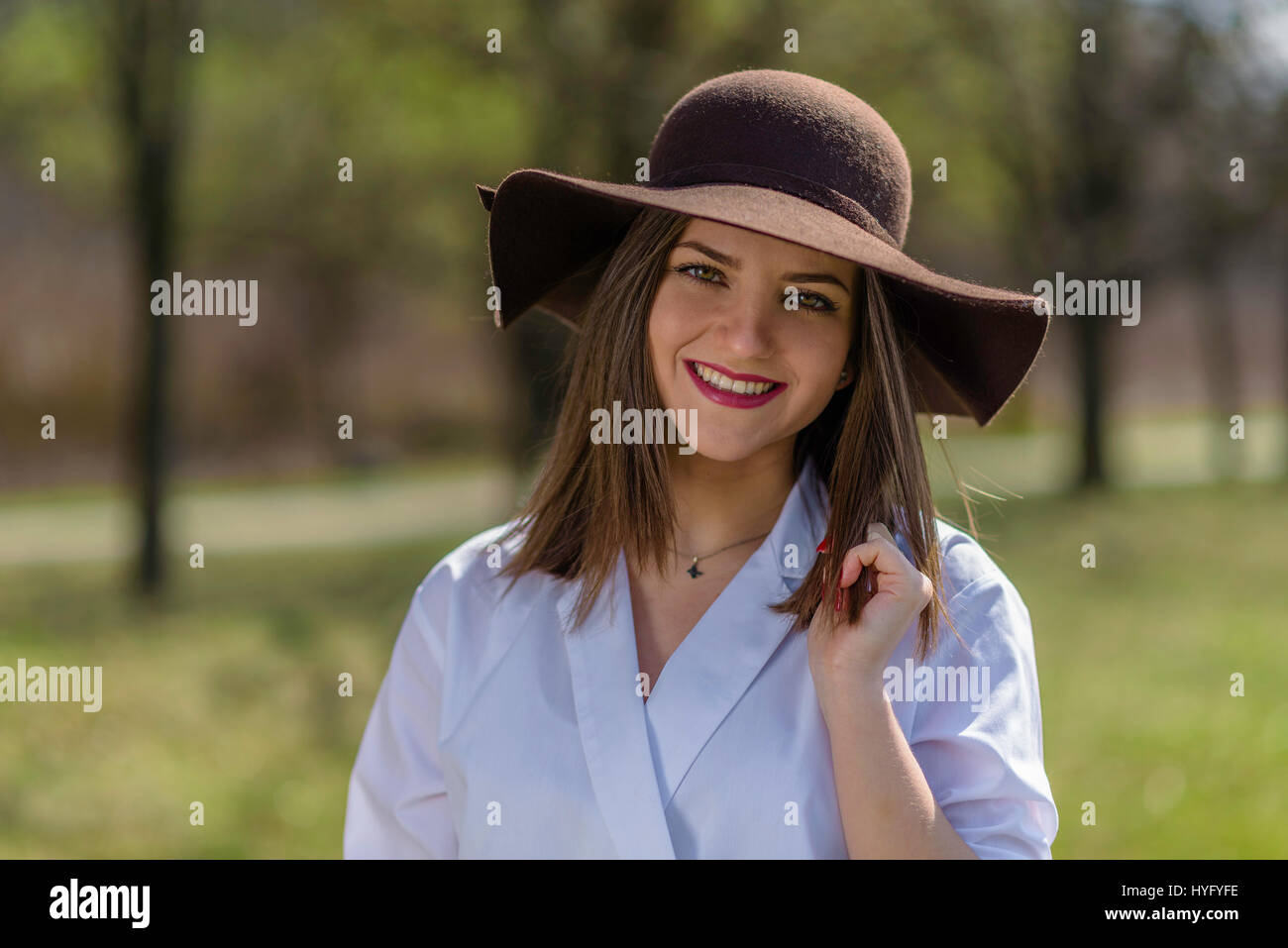 Portrait of a smiling young woman wearing a hat in a park during spring. Woman bowed her head head to the right side of frame. Medium shot. Shallow de Stock Photo