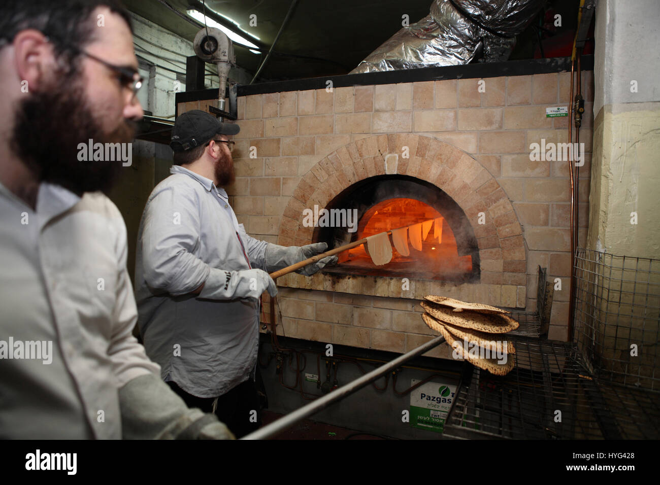 One Hasidic man puts dough pieces in the oven, the other one, unseen, takes the ready Matza out, both use long sticks and are in a handmade factory. Stock Photo