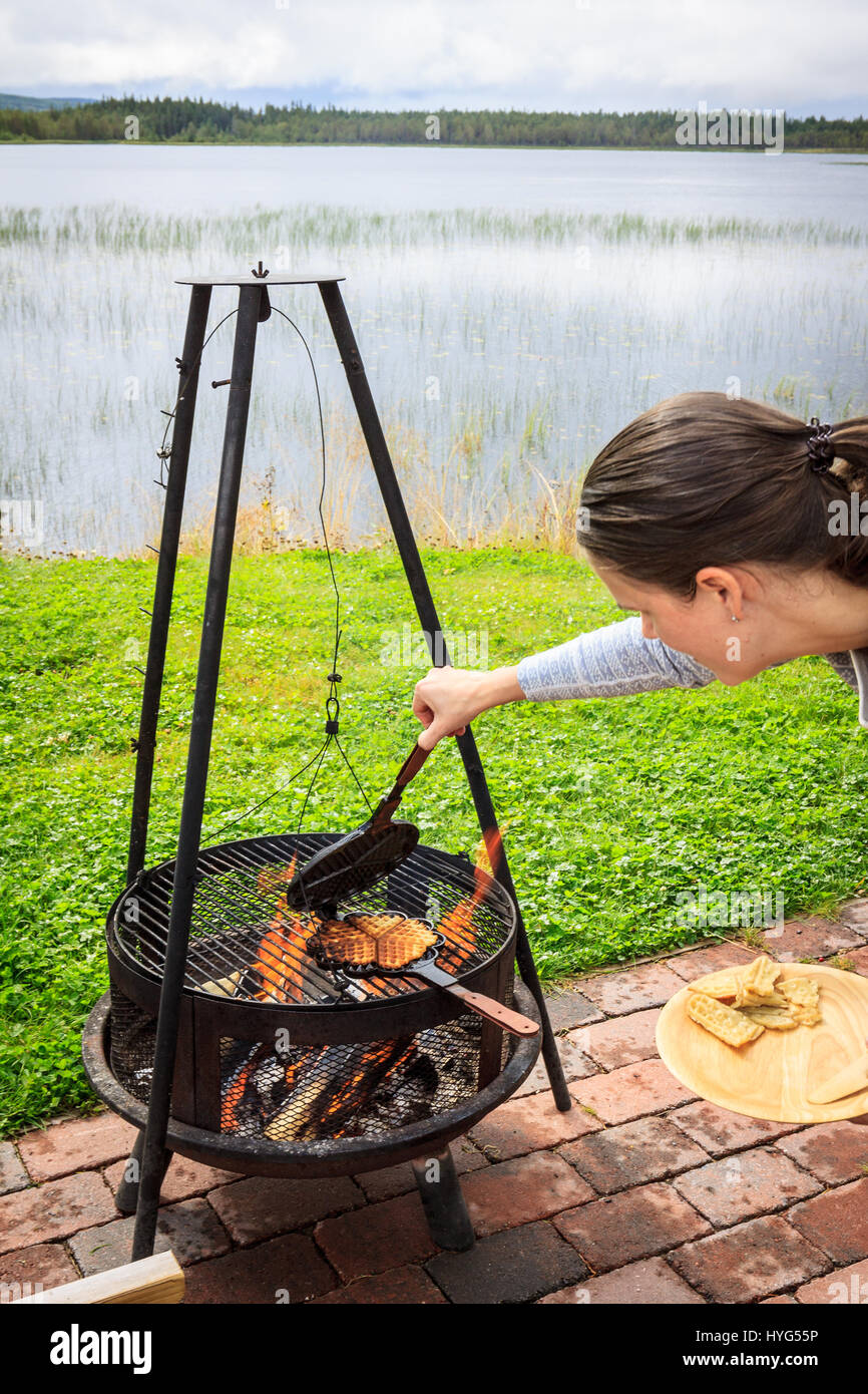 Young woman checking cast-iron waffle iron on camp fire, with freshly baked waffle inside. Camping in nature, next to calm lake. Stock Photo