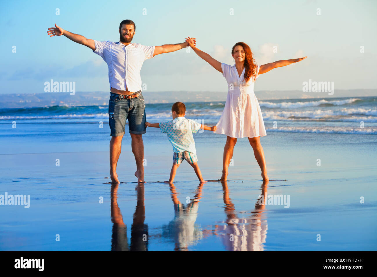 Happy family - father, mother, baby son hold hands, walk and jump together by water pool on sunset black sand beach with sea surf. Travel, active life Stock Photo