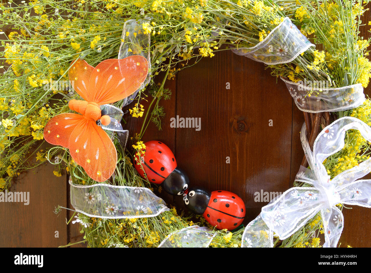 Wreath of yellow wild flowers and decorated with an orange butterfly and two wooden ladybugs on wooden background Stock Photo