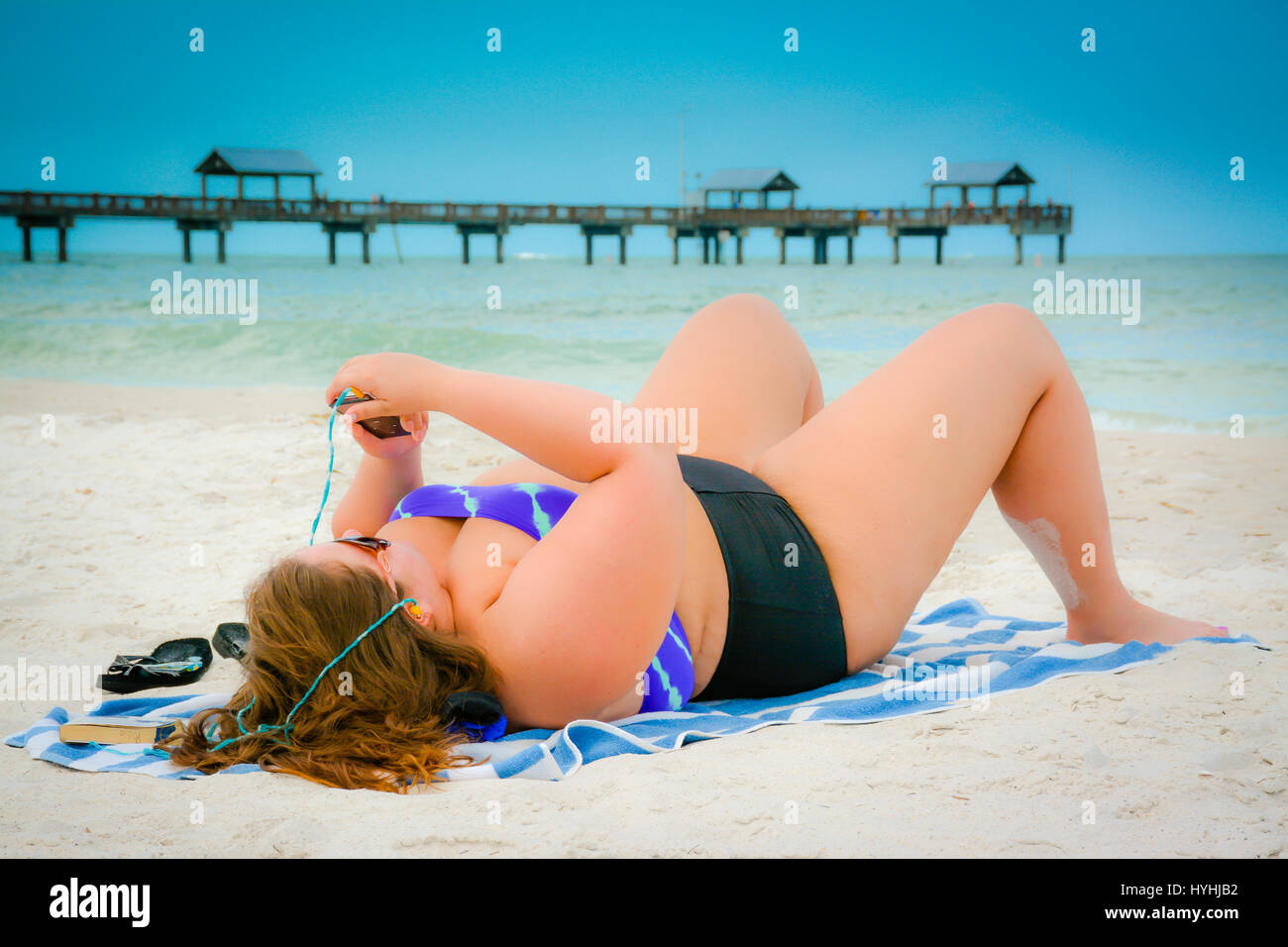 Plus size woman in two piece swimsuit lying on Clearwater Beach, FL holding Smart phone with earphones and Pier 60 in the Gulf of Mexico Stock Photo