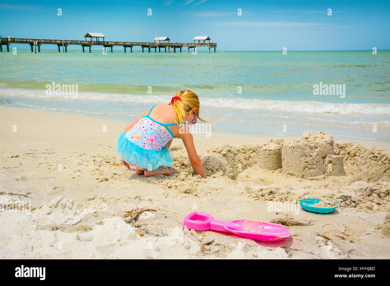 Rear view of young blond girl in blue bathingsuit plays on white sand beach at Clearwater Beach, FL with Pier 60 and the shoreline nearby on the Gulf  Stock Photo