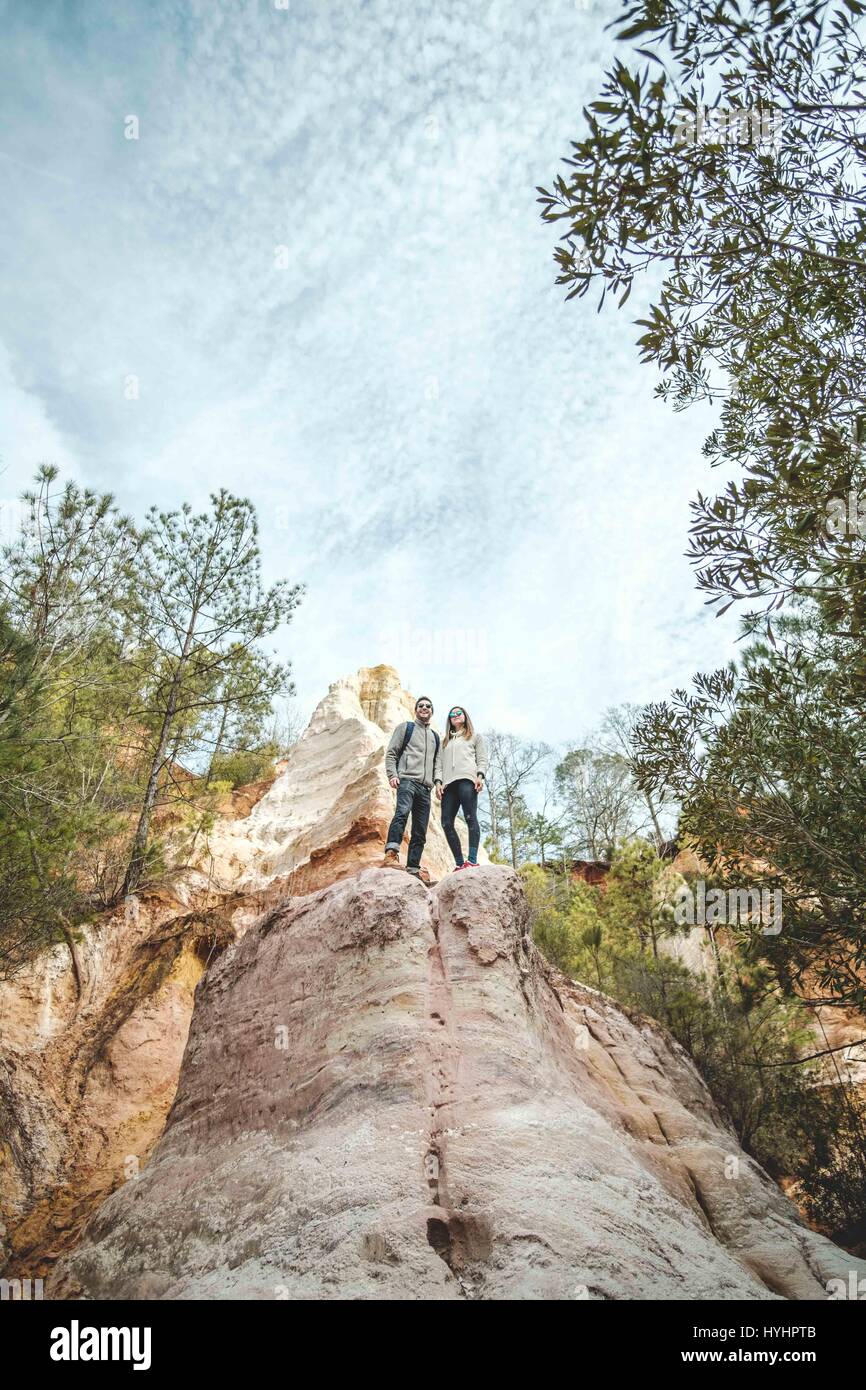 Portrait of a young couple from a low perspective standing on a rock formation with trees and the sky. Stock Photo