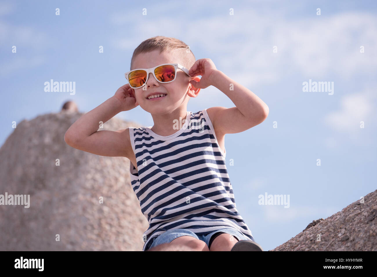 happy little boy in a vest and sunglasses sitting on breakwater block on the background of the sky Stock Photo