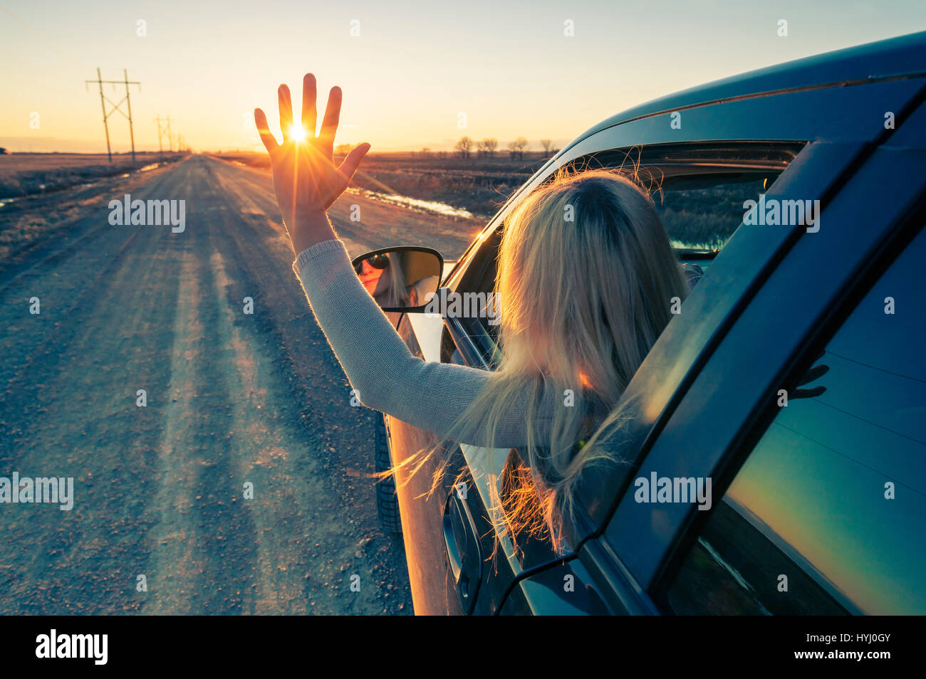 Woman driver is waving to the sun at sunset on the road. Stock Photo