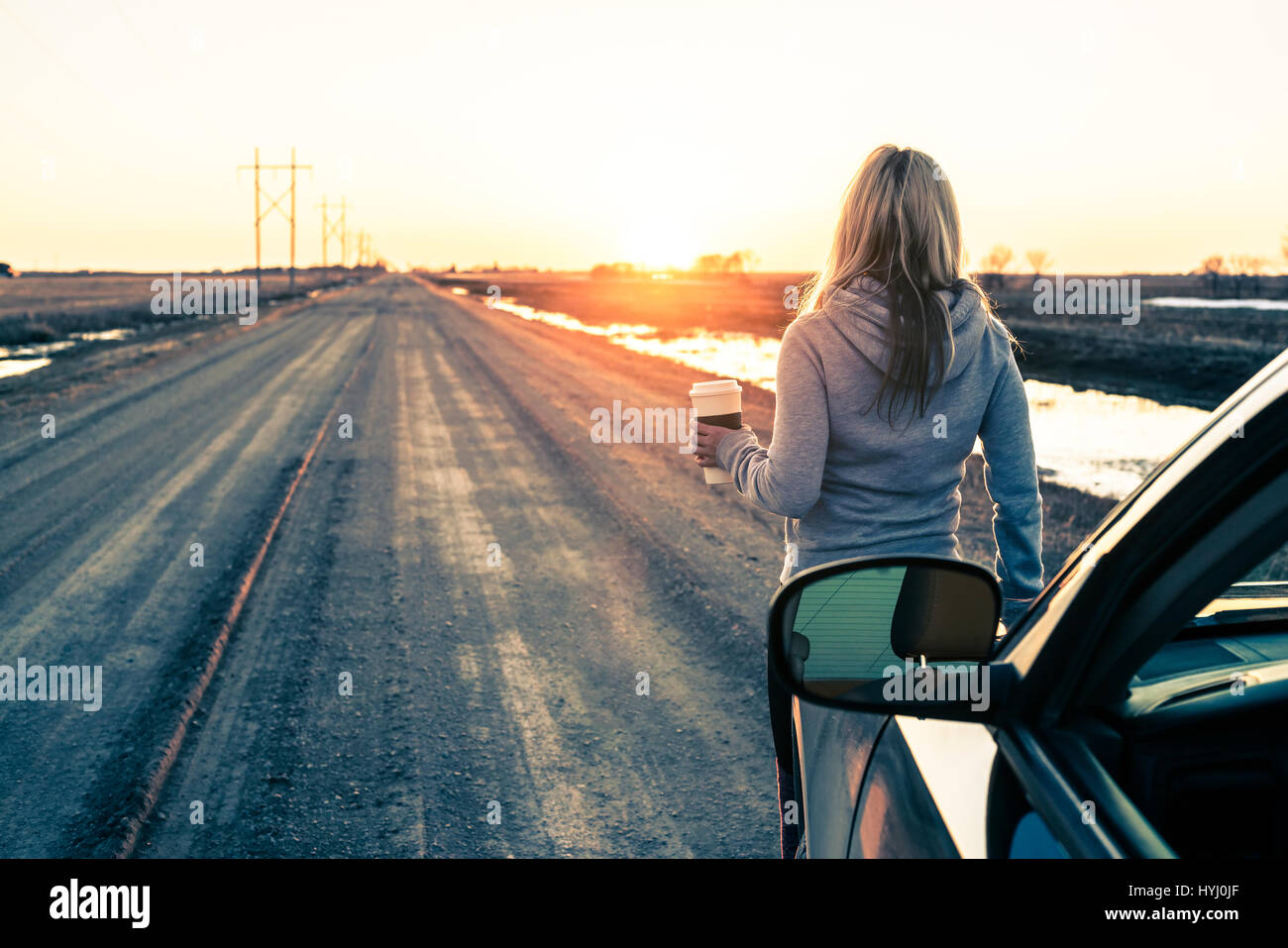 Woman standing with her car, holding caffe cup and watching sunset on the road Stock Photo