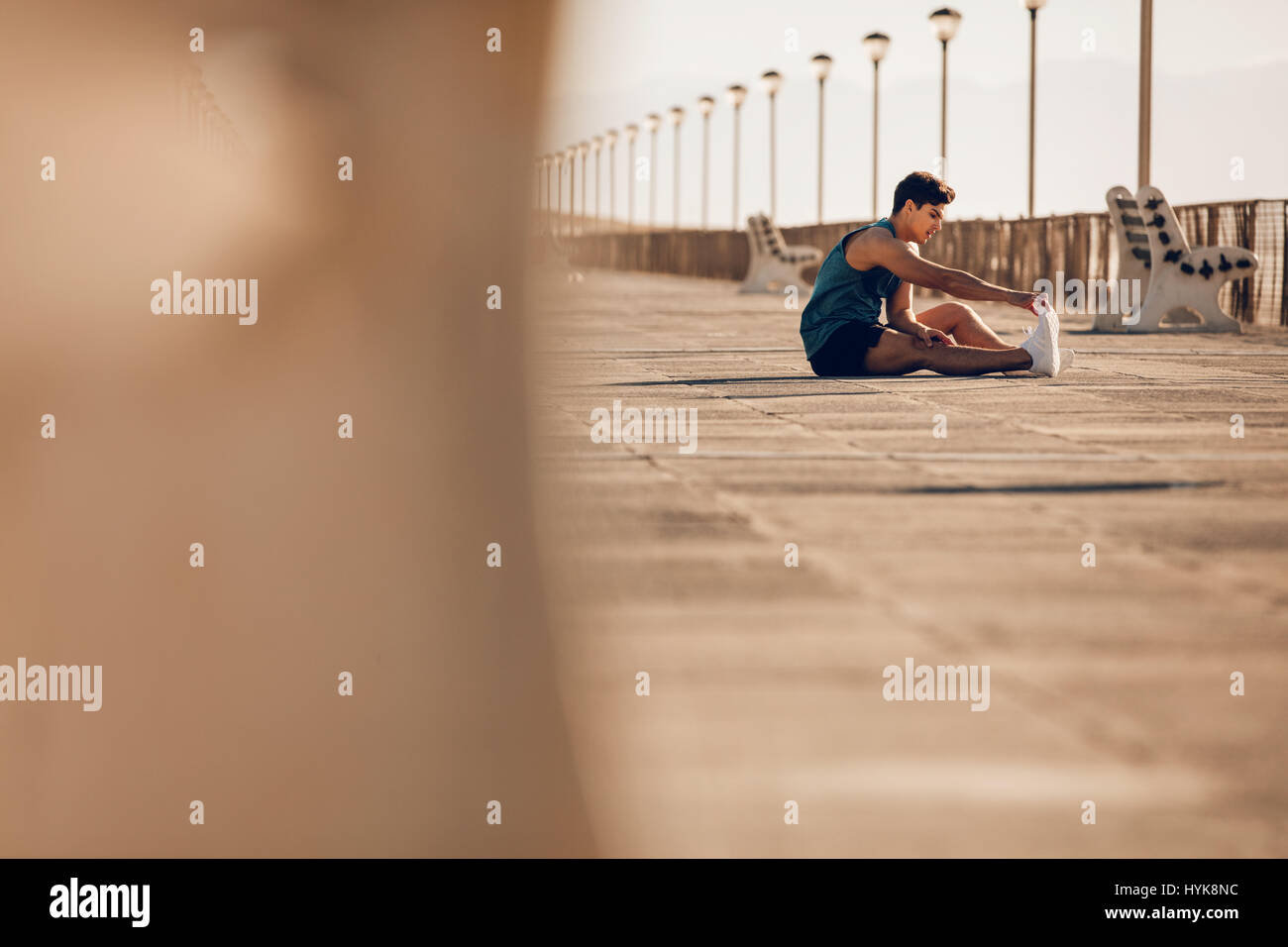 Shot of healthy young man taking a break running session. Male runner stretching legs after running on promenade. Stock Photo