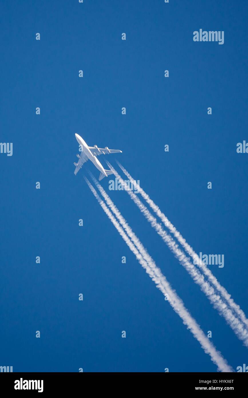 Vertical photo of big white ariplane with four engines taken from the ground. The strong white smoke is visible how is created on clear dark blue sky  Stock Photo