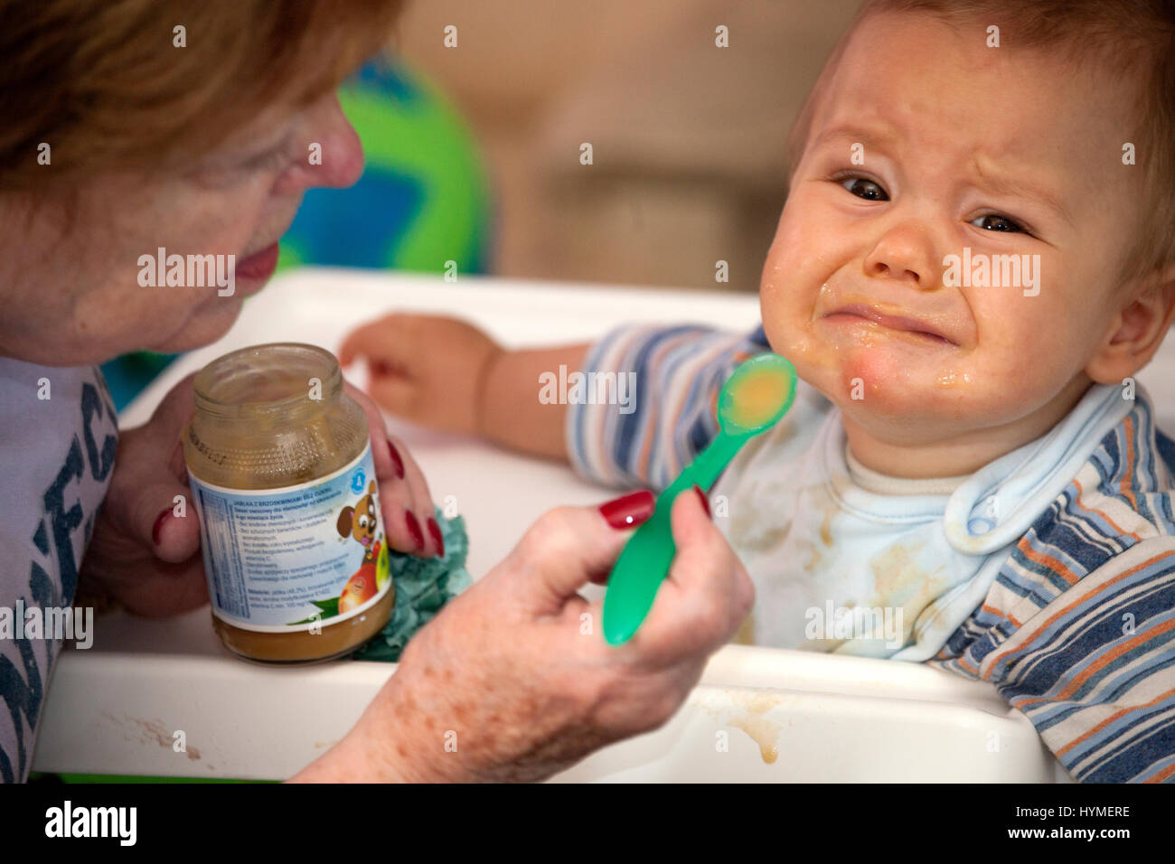 Grandmother trying to feed very unhappy grandson. Grandma age 74 and Polish baby 7 months old. Zawady Central Poland Europe Stock Photo