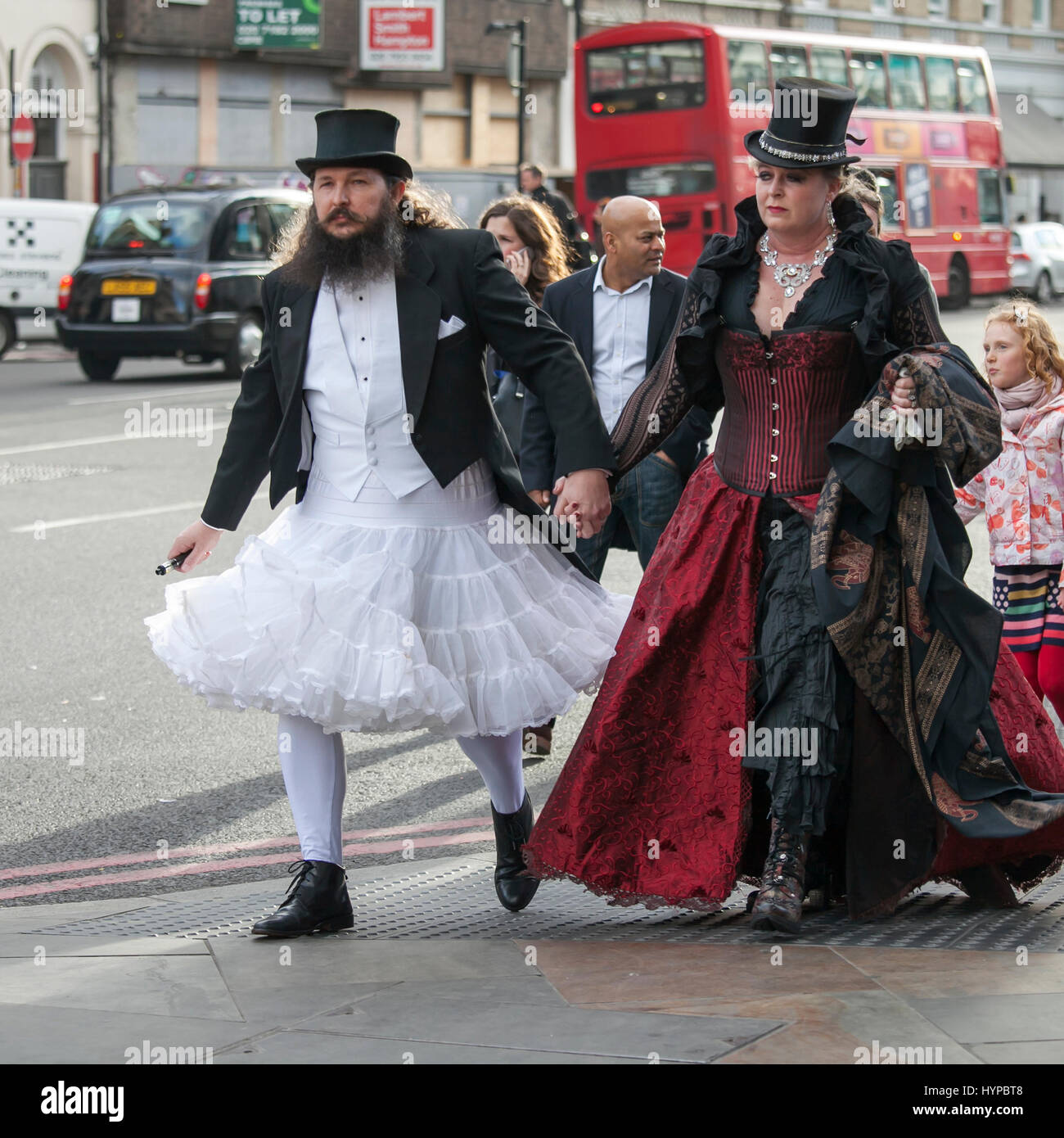 LONDON, ENGLAND - March 30, 2017 A man in a white wedding dress and a top hat with a woman in a gothic red velvet dress walking down the street in Lon Stock Photo