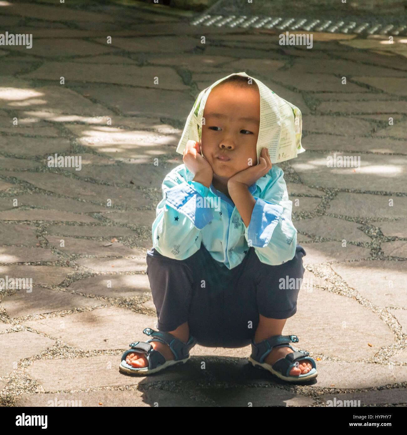 Playful young Chinese boy sitting on a path wearing newspaper hat Stock Photo