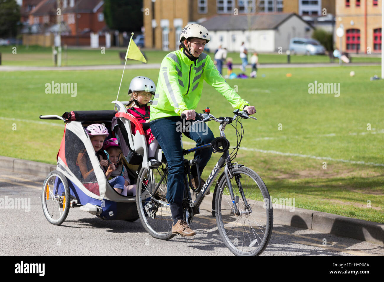 Woman cyclist on bike / bicycle with + 3 children; Co Pilot child seat with helmet & towing cycle Chariot trailer with two / 2 kids with helmets. UK. Stock Photo