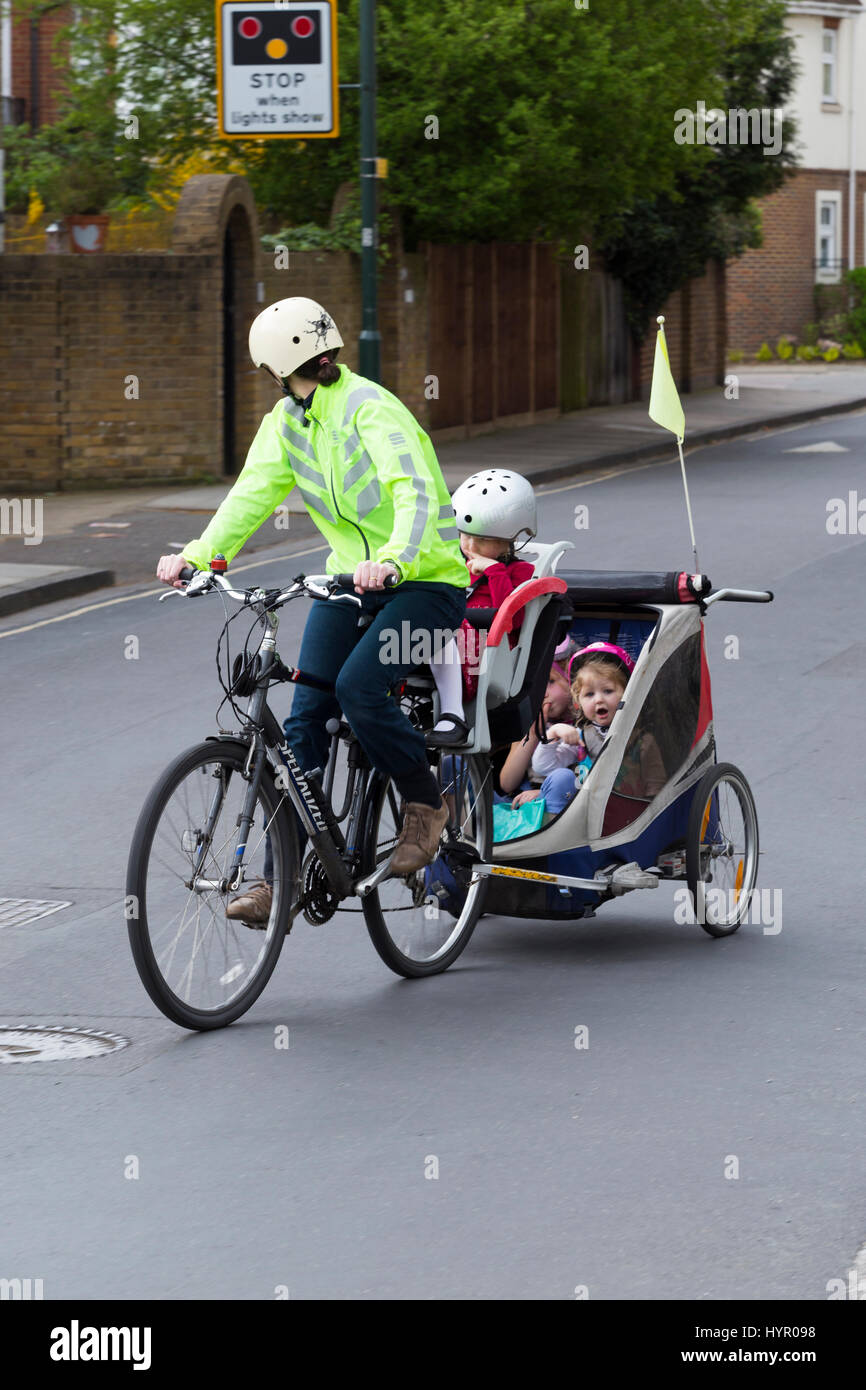 Woman cyclist on bike / bicycle with + 3 children; Co Pilot child seat with helmet & towing cycle Chariot trailer with two / 2 kids with helmets. UK. Stock Photo