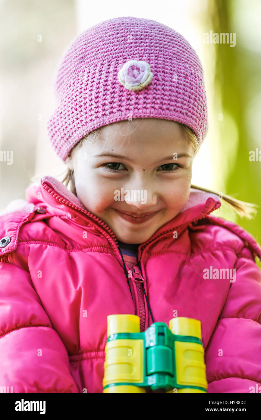 Portrait of a little girl in pink with binoculars around her neck, exploring nature, being impish and timid for the camera. Active outdoor lifestyle c Stock Photo