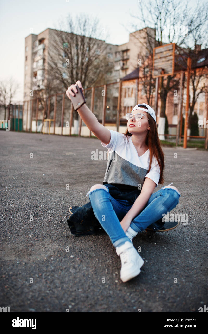 Young teenage urban girl with skateboard, wear on glasses, cap and ripped  jeans at the yard sports ground on sunset making selfie on phone Stock  Photo - Alamy