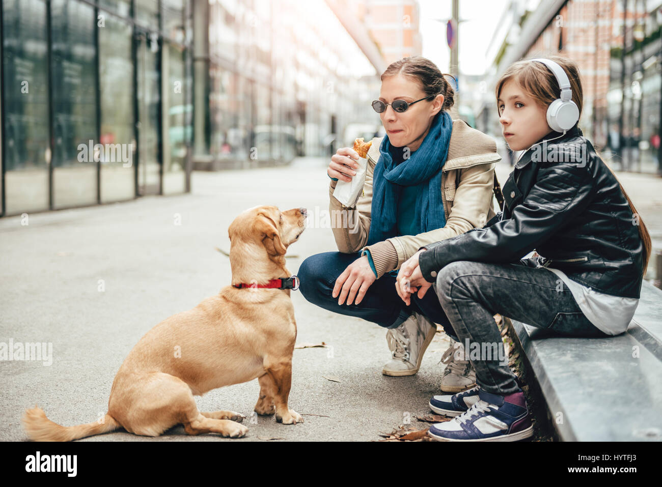 Mother eating sandwich and looking at a dog while daughter listening to music on the street Stock Photo