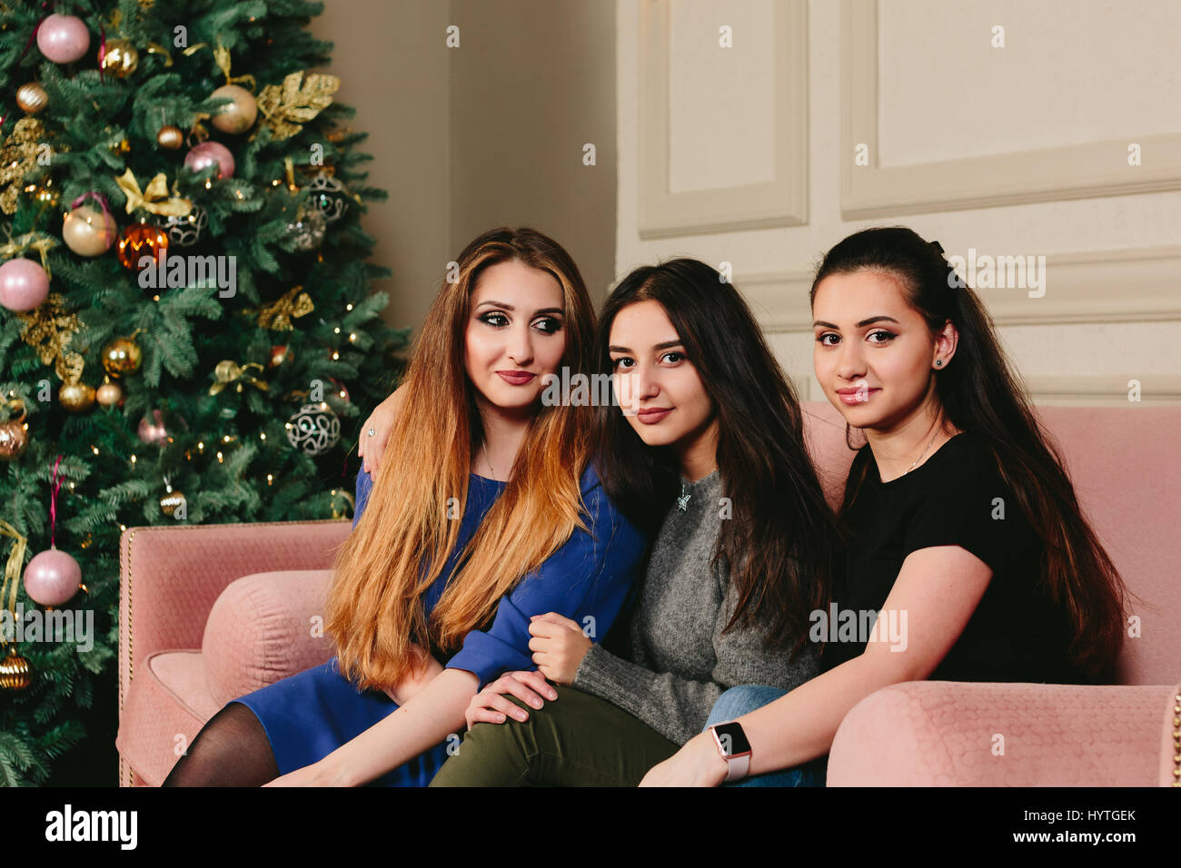 Three beautiful young girlfriends on a sofa near the Christmas tree. Studio horizontal portrait. Stock Photo
