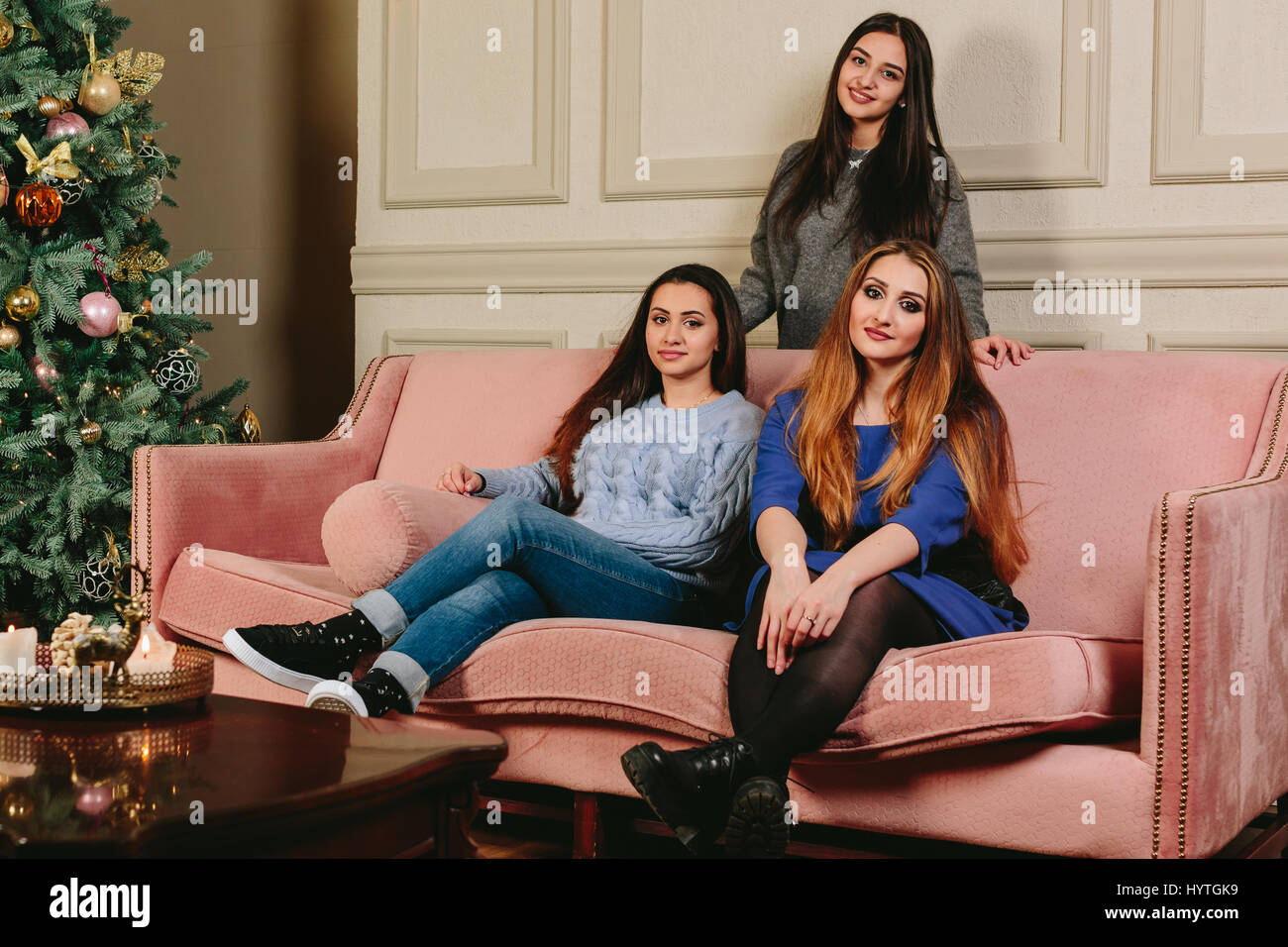 Three beautiful young girlfriends on a sofa near the Christmas tree. Studio horizontal portrait. Stock Photo