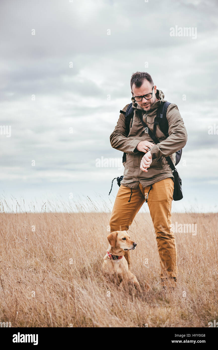 Hiker with dog standing in grassland and looking at watch Stock Photo