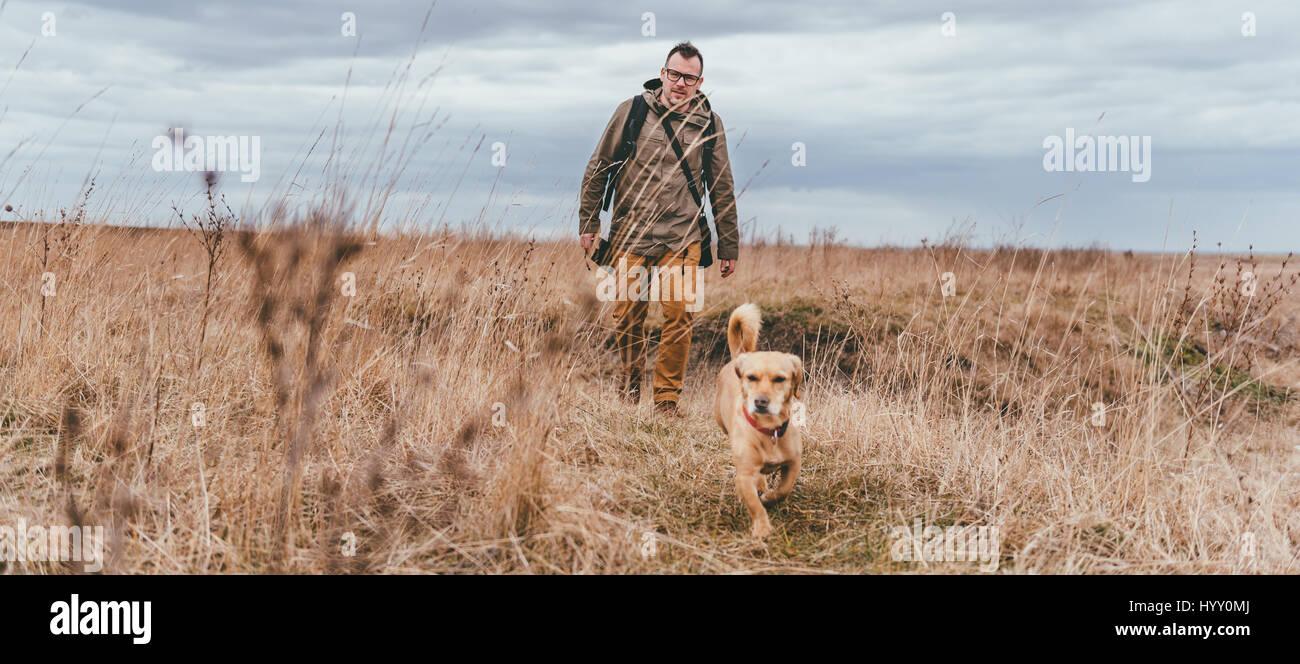 Hiker and small yellow dog walking in grassland on a cloudy day Stock Photo