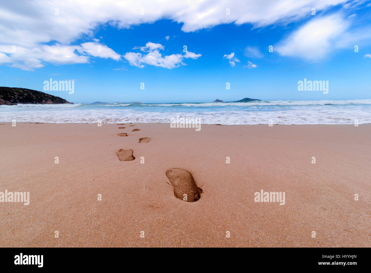 A low angle view of a sandy beach with foot prints leading towards the waves with a blue sky leading towards the horizon. Stepping into the horizon. Stock Photo