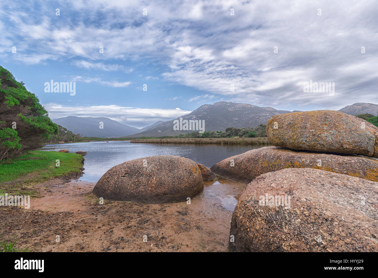 A beautiful landscape view of a distant mountain range with a tree line and large rocks in the foreground and a clam river flowing down the middle. Stock Photo