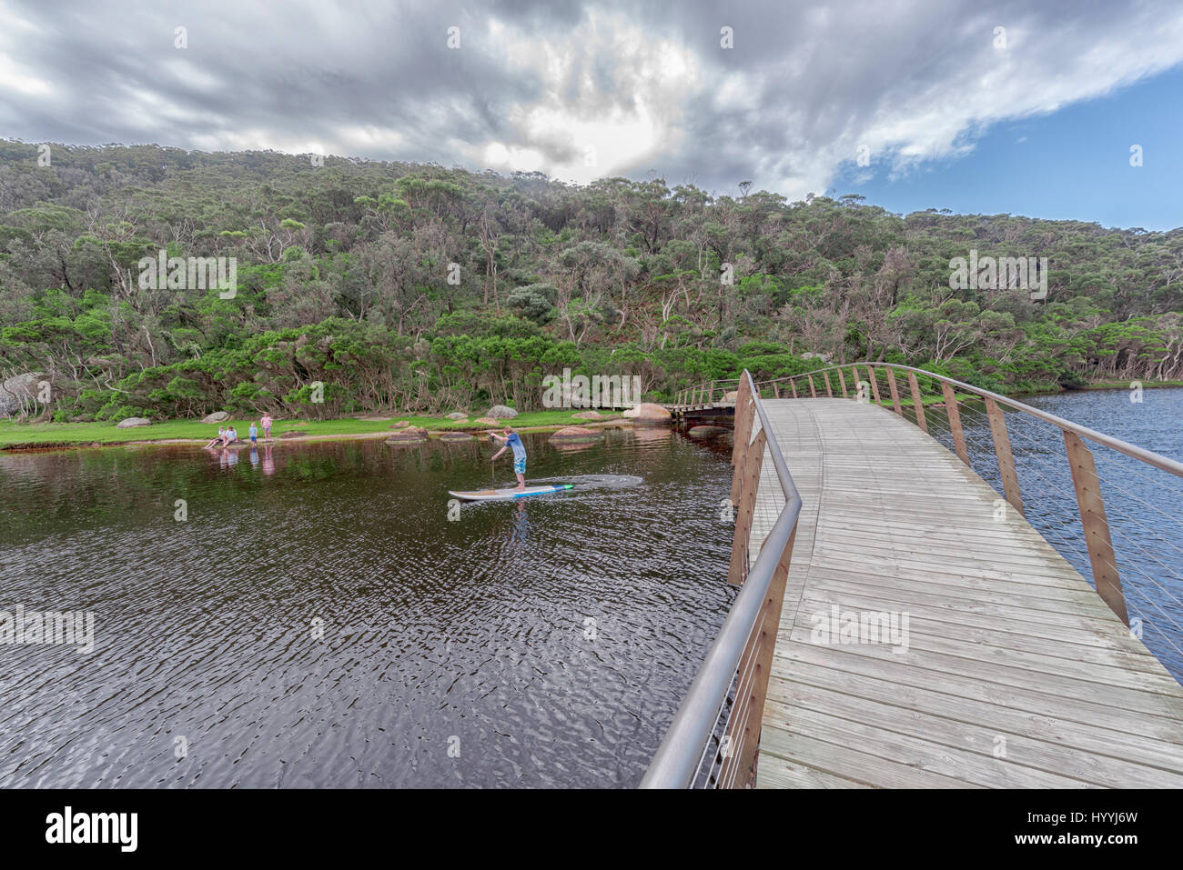 Small bridge leading to a forested area over a river with kids playing on the beach in the distance and a paddle boarder on the river Stock Photo