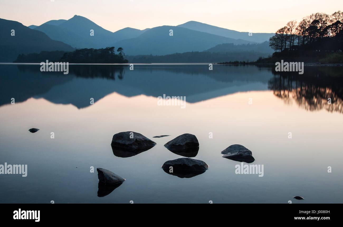 Rocks and the mountains of the Derwent Fells are reflected in the calm waters of Derwent Water lake in England's Lake District National Park. Stock Photo