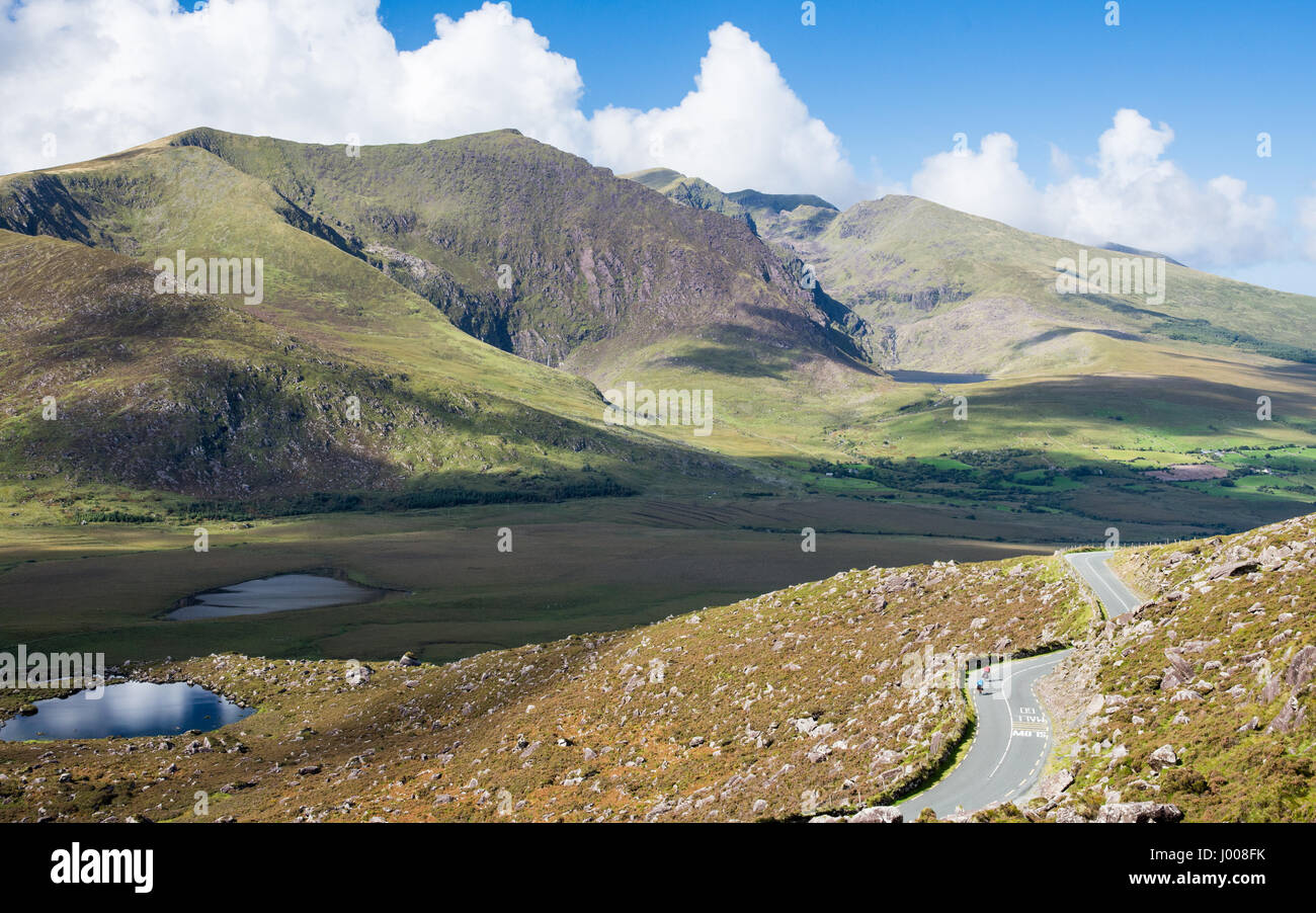 Cyclists descend the steep Conor Pass road through the mountains of Ireland's Dingle Peninsula, with the sun lit slopes of Brandon Mountain behind. Stock Photo