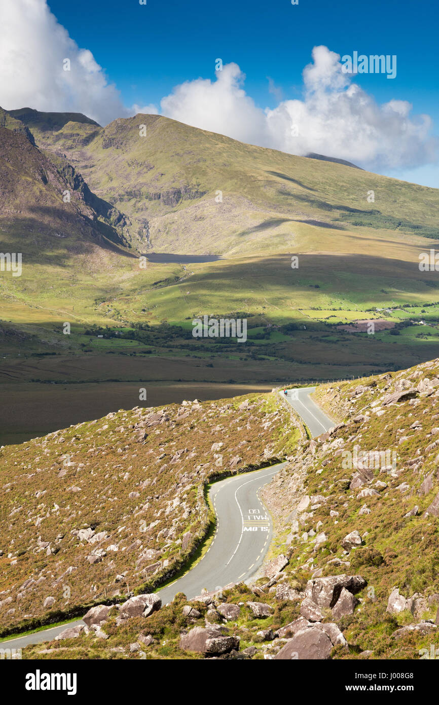 Cyclists descend the steep Conor Pass road through the mountains of Ireland's Dingle Peninsula, with the sun lit slopes of Brandon Mountain behind. Stock Photo