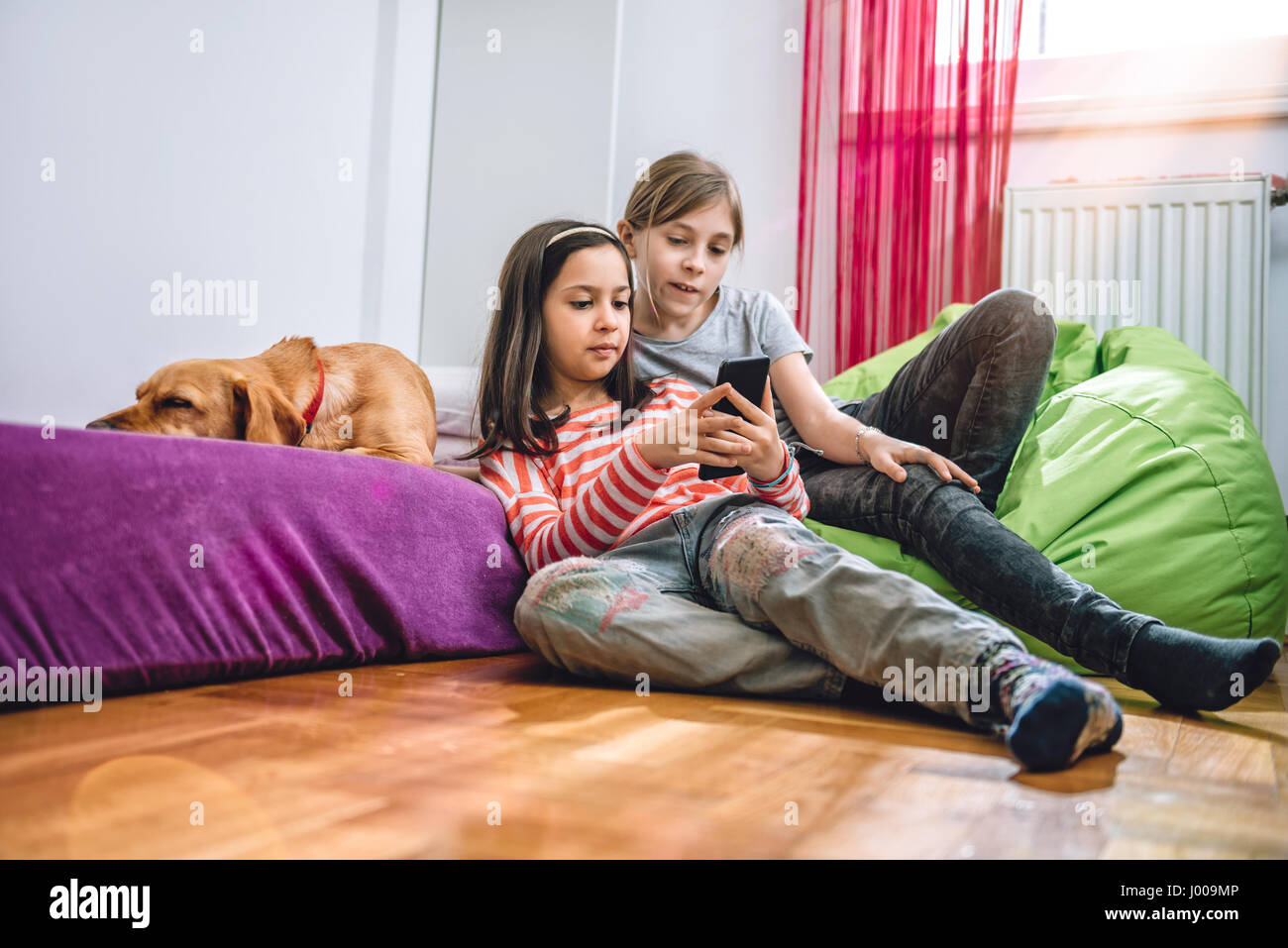 Two girls with small yellow dog sitting in the kids room and using smart phone Stock Photo
