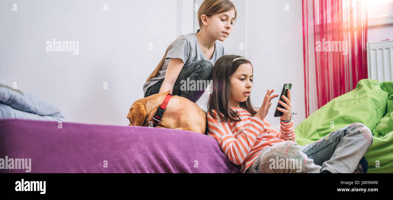 Two girls with small yellow dog sitting in the kids room and using smart phone Stock Photo