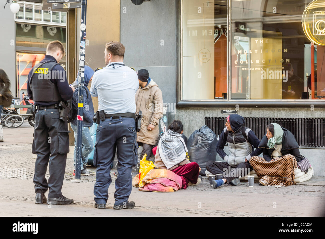 Two policemen looking at the documents of some immigrants, who are sitting on the street. Copenhagen, Denmark - April 6, 2017 Stock Photo