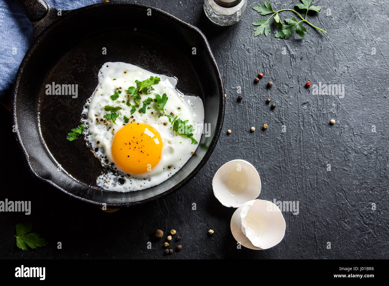 Fried egg. Close up view of the fried egg on a frying pan. Salted and spiced fried egg with parsley on cast iron pan and black background. Stock Photo
