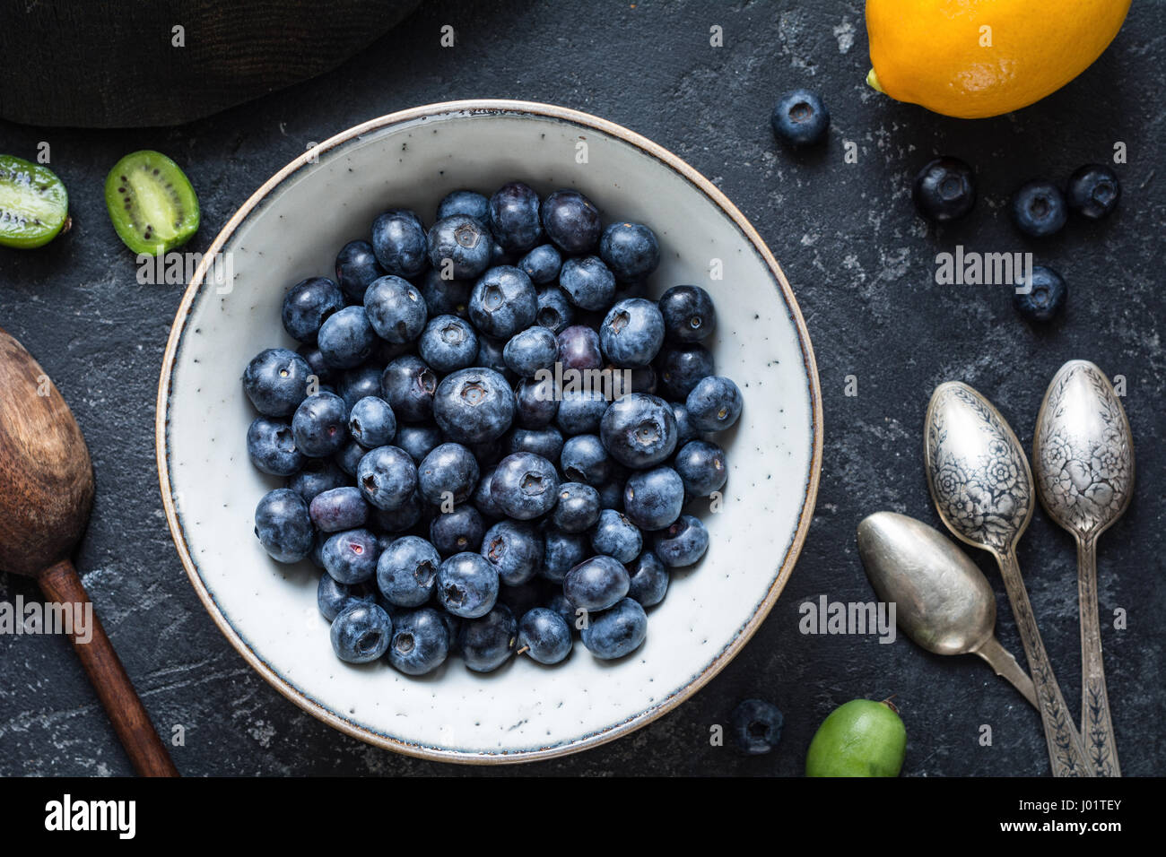 Blueberries and vintage spoons: Top view of fresh blueberries in white ceramic bowl with baby kiwi fruit, lemon and vintage tea spoons on dark stone t Stock Photo