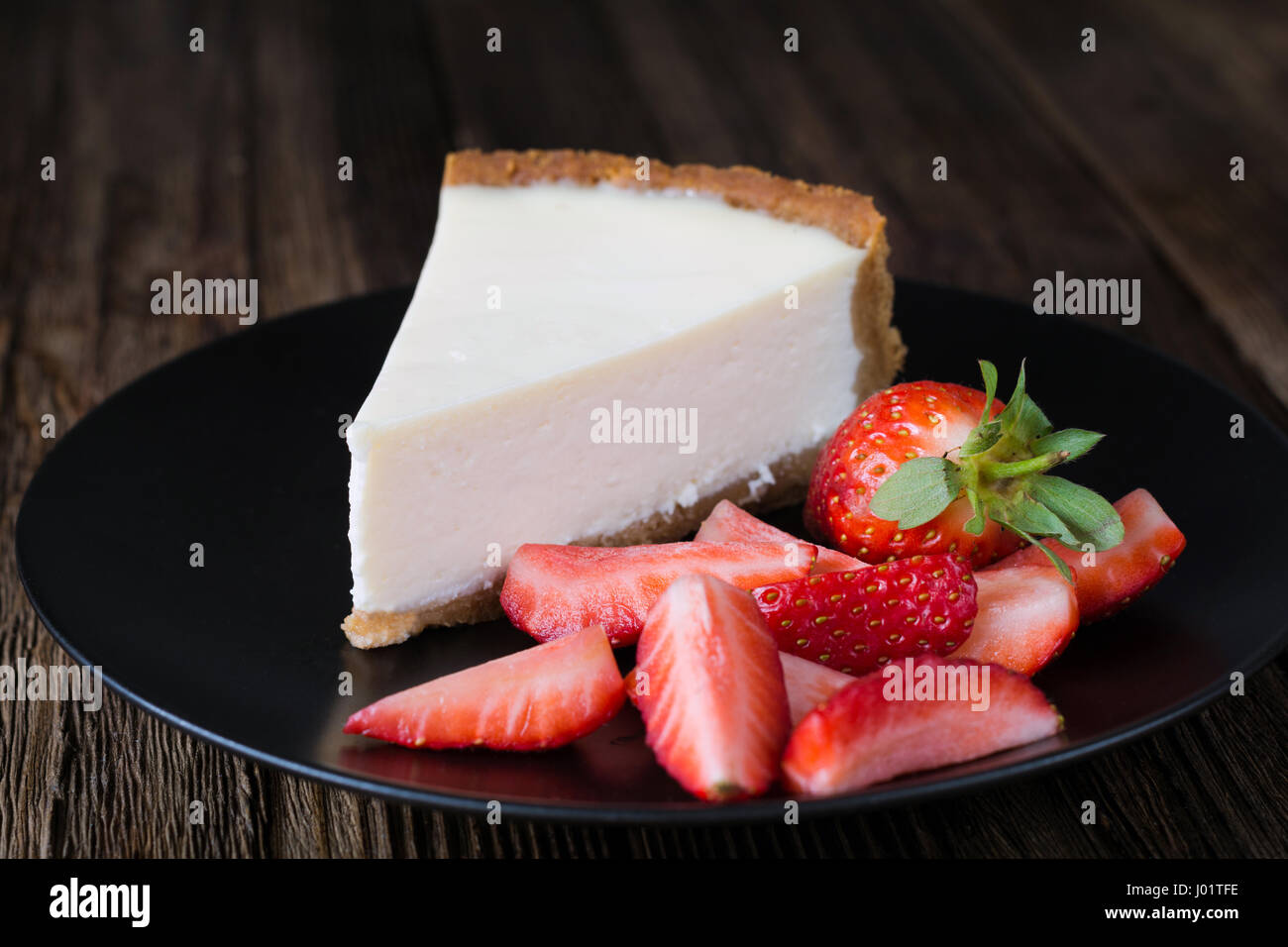 Cheesecake with fresh strawberries on black plate. Slice of plain cheesecake. Wooden table background Stock Photo