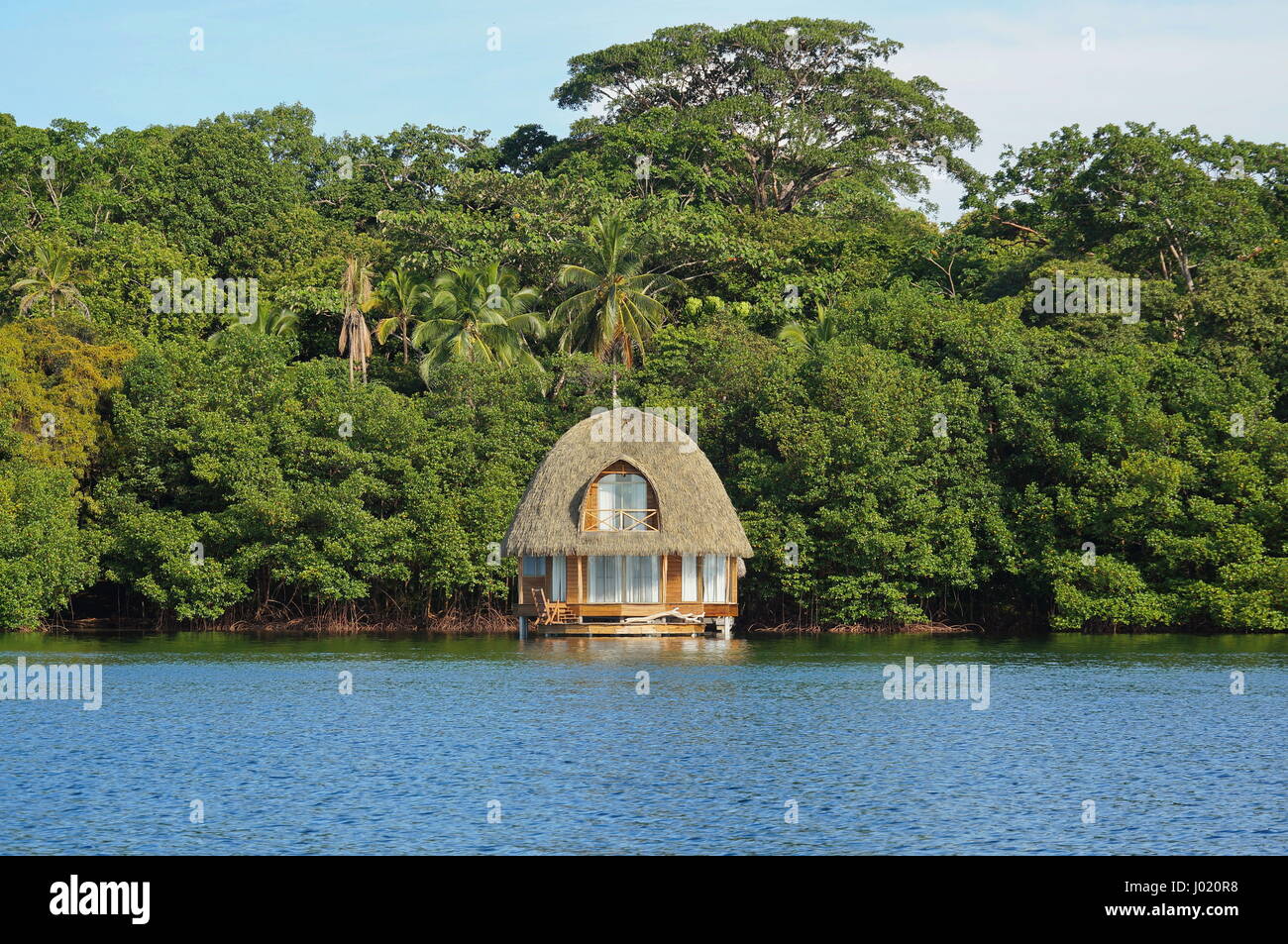 Tropical bungalow overwater with thatched palm roof and lush green vegetation on the sea shore, Bocas del Toro, Caribbean, Central America, Panama Stock Photo