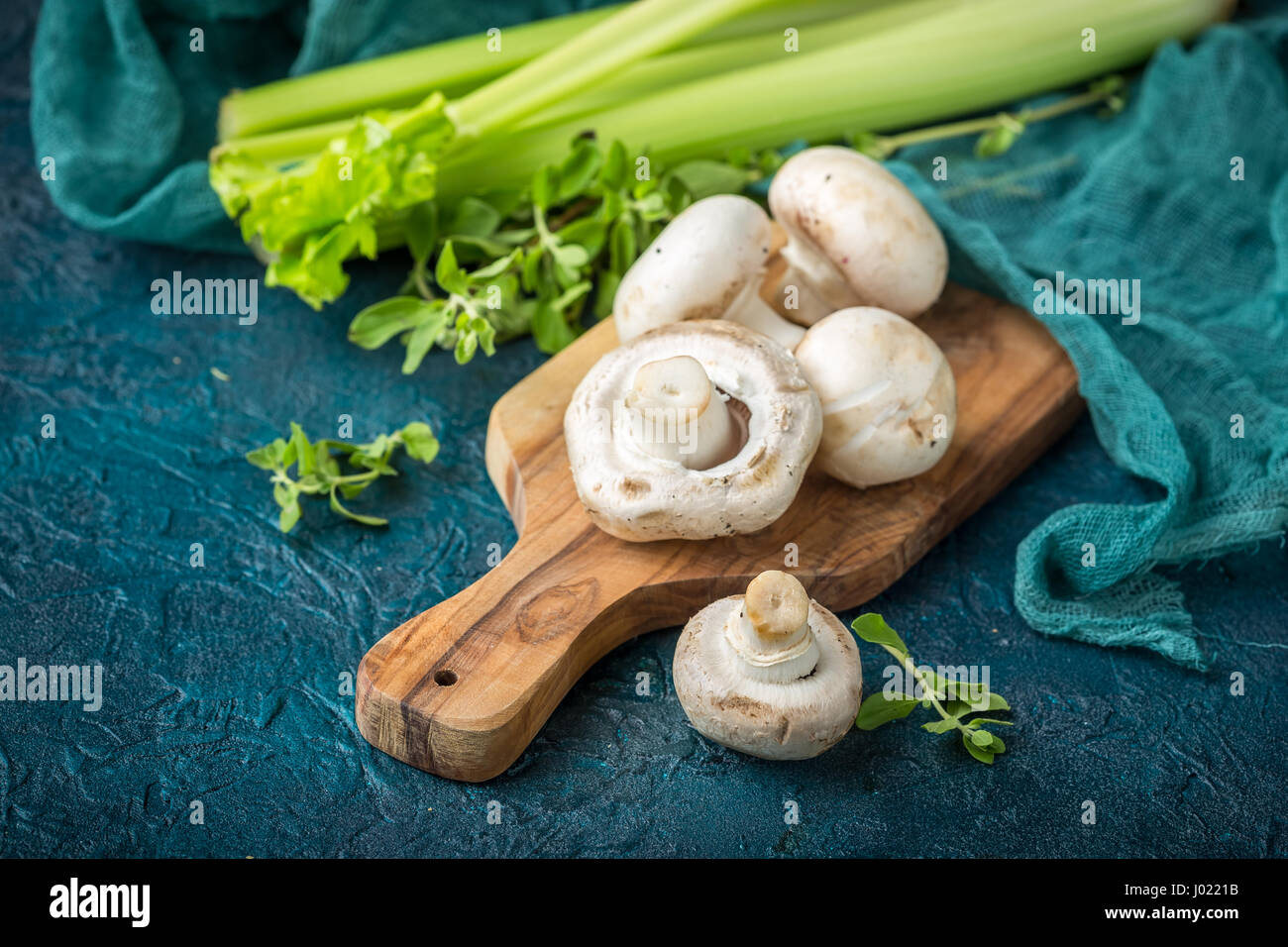 Fresh mushrooms champignons on a wooden cutting board, parsley and stalks of celery on a dark blue background Stock Photo