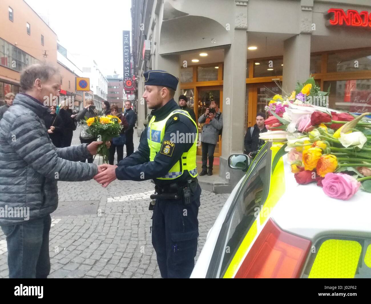 Swedish citizen expresses his condolences by giving a bouquet of flowers to a (Polis) police officer sitting in front of a parked police car covered by flowers deposited by pedestrians in memory to the victiims of the terrorist truck attack in the center of Stockholm's  city centre. Stock Photo
