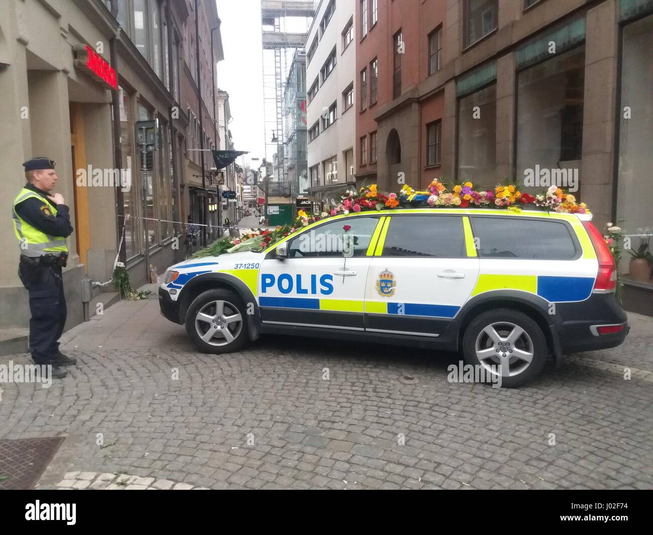 Swedish Polis policeman standing sideways to a police car parked in a central street in Stockholm, covered by a pile of hundreds of flower bouquets, symbolising the generous Swedish sympathy and mourn for the lost lives of their compatriots, after the terrorist attack led by man driving a truck in high speed, slamming into pedestrians around Ahlens shopping centre. One man has been arrested insofar, after a manhunt  for the suspected attacker who left 4 dead and 16 injured civilians behind his actions. Stock Photo