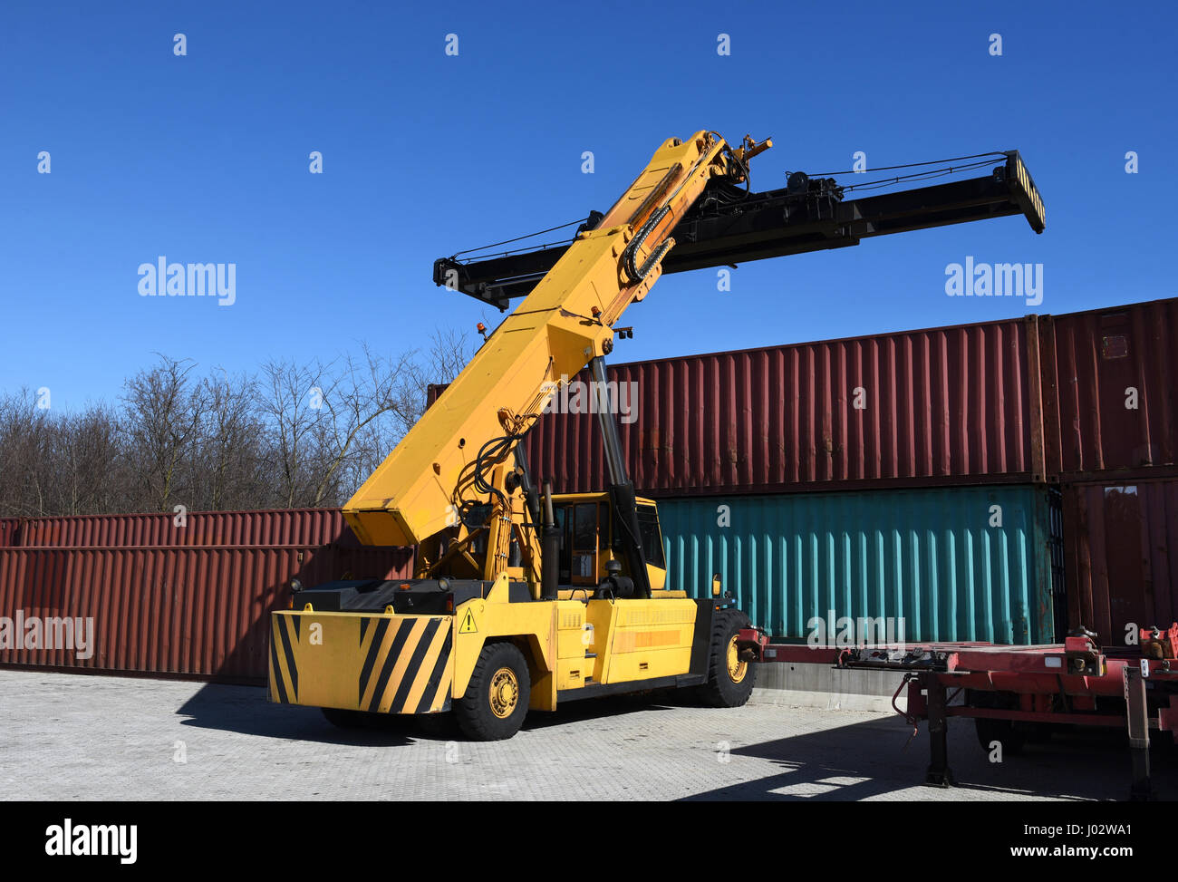 Yellow container handling vehicle in action near stack of metal containers outdoors view from the back Stock Photo
