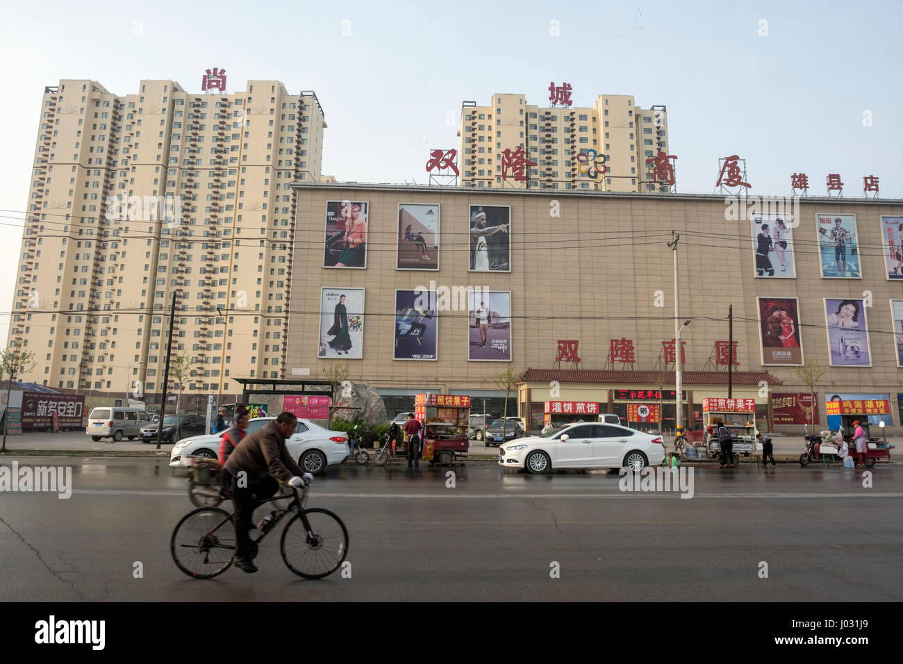 A man rides a bicycle past a department store and a compound of apartments in Xiong County in Baoding, north China's Hebei province. 09-Apr-2017 Stock Photo