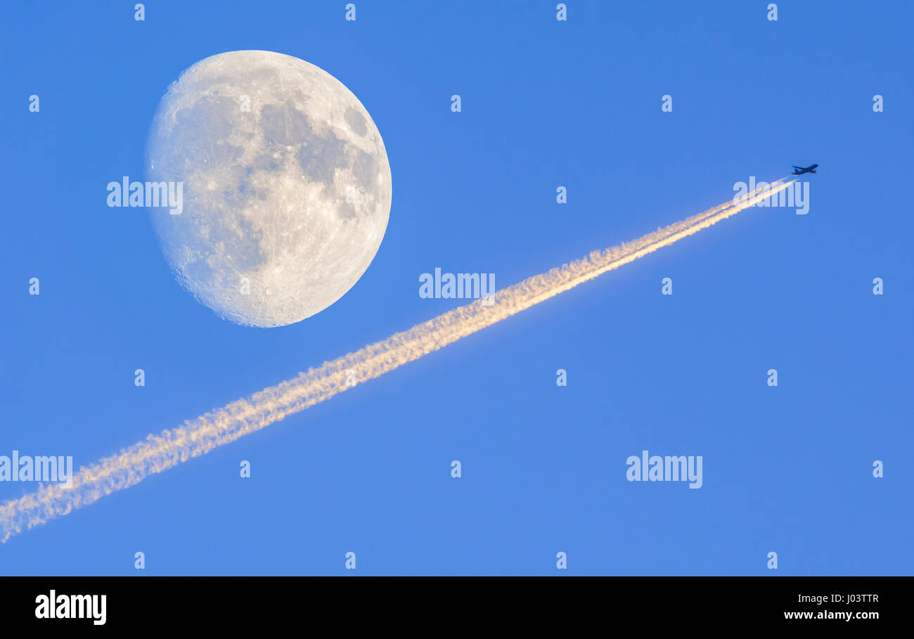 Condensation trails (contrails) from a jet plane with a nearly full moon (at waxing gibbous), against blue sky. Stock Photo